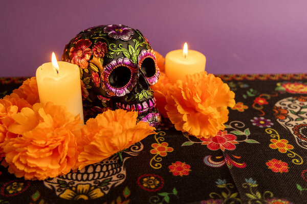 Череп украшен металлическими цветами, рядом с ним мерцают две свечи. Сцена окружена оранжевыми цветами, так как вокруг аккуратно расставлено больше свечей.