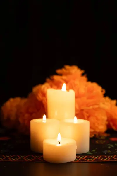 stock image A vertical view of three lit candles encircled by vivid orange marigold flowers on a Day of the Dead themed tablecloth. The black background emphasizes the soft candlelight, creating a serene scene.