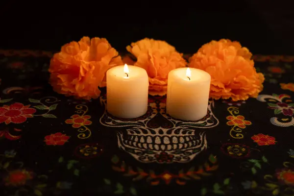 stock image A dark background highlights three lit candles surrounded by vibrant orange marigold flowers. The candles warm glow contrasts with the Dia de los Muertos patterned tablecloth, creating a serene atmosphere.