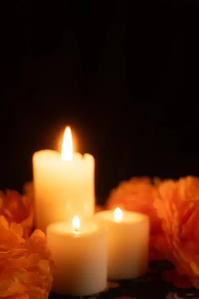 Stock image Three lit candles cast a warm glow from the left, surrounded by orange marigold flowers. The scene is set on a Day of the Dead tablecloth, against a deep black background, capturing the essence of the celebration.