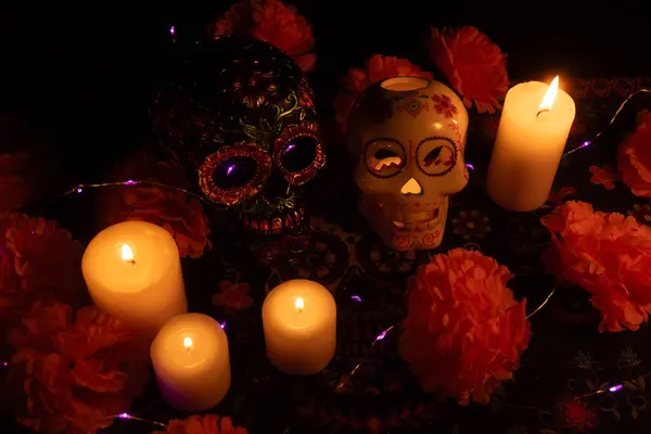 stock image A vivid Dia de los Muertos scene featuring two skulls: one adorned with metallic flowers and another hollow with a lit candle inside. Surrounded by marigold flowers and candles on a decorated tablecloth, illuminated by candlelight against a black ba