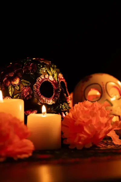 Stock image A vertical image showing two skulls with distinct decorations on a Day of the Dead cloth. One skull is adorned with metallic flowers while the other has a lit candle inside. Surrounded by marigolds and candles.