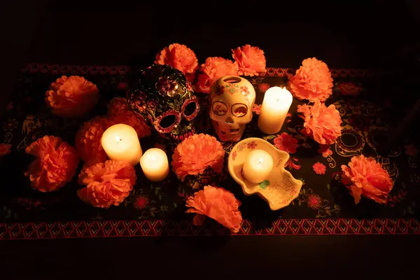 stock image A general view featuring two skulls on a Day of the Dead themed tablecloth. One skull is adorned with metallic flowers, the other hollow with a lit candle inside. Surrounded by marigold flowers and lit candles.