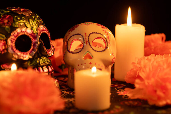 Поразительное зрелище с металлически-цветочным украшенным черепом и полым черепом с зажженной свечой, установленным в день Мертвых тканей. Оранжевые ноготки и свечи окружают центральную часть, все на черном фоне.
