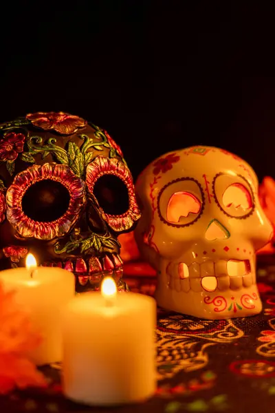 stock image Vertical composition featuring two skulls on a Day of the Dead tablecloth: one decorated with metallic flowers, the other hollow with a lit candle. Marigold flowers and additional candles create a festive atmosphere against a black background.