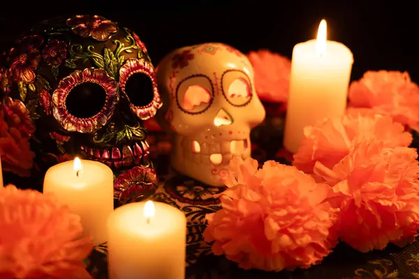 stock image Two decorated skulls sit on a Dia de los Muertos tablecloth: one adorned with metal flowers and the other with a lit candle. Marigold flowers and surrounding candles create a traditional Mexican festival scene.