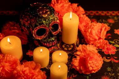 Close-up of a skull decorated with metallic floral accents, encircled by orange marigold flowers. Five candles light the scene, with a Day of the Dead motif tablecloth and a black background enhancing the festive atmosphere.