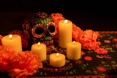 Metalik çiçeklerle süslenmiş ve canlı turuncu kadife çiçekleriyle çevrili detaylı bir kafatası. Etrafına beş mum konur, hepsi de siyah bir arkaplan üzerine kuruludur, Dia de los Muertos 'un özünü yakalar..