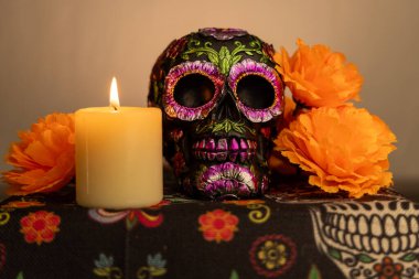 Metalik çiçeklerle süslenmiş detaylı bir kafatası canlı turuncu kadife çiçekleriyle çevrilidir. Aydınlatılmış bir mum, beyaz bir gradyan zemin üzerine sıcaklık ekler, Dia de los Muertos temaları için mükemmeldir..