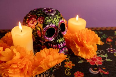 An artistic skull with metallic floral decorations, flanked by two burning candles. Orange flowers and additional lit candles create a warm, festive atmosphere.