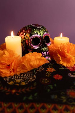 Metalik çiçeklerle süslenmiş bir kafatası canlı turuncu çiçeklerle çevrili iki mum arasında oturur. Dikey çekim Dia de los Muertos atmosferini yakalar ek mumlar yerleştirilir.