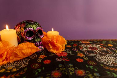 A skull decorated with metallic flowers, lit by two candles on each side. Orange flowers encircle the scene, with more candles being carefully arranged, symbolizing Dia de los Muertos. clipart