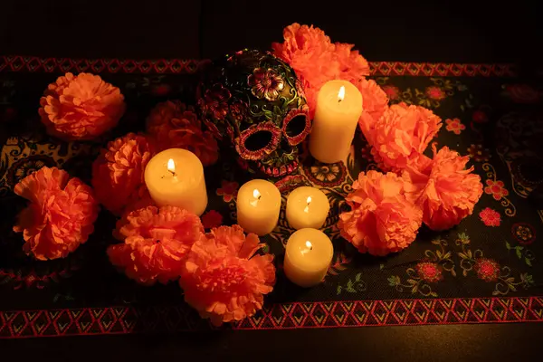 stock image Top view of a decorated skull adorned with metallic flowers, surrounded by vibrant orange marigold petals. Lit candles and a Day of the Dead patterned tablecloth enhance the traditional Mexican celebration.