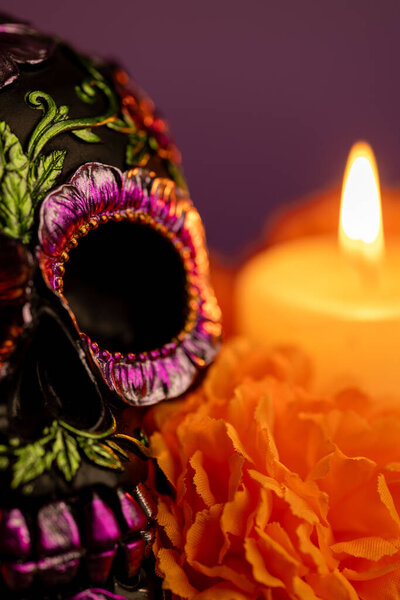 Вертикальный крупный план детального черепа, украшенного металлическими цветами, окруженный яркими оранжевыми цветами. Рядом с ним зажжена свеча, которая освещает празднование Дня мертвых в Мексике.