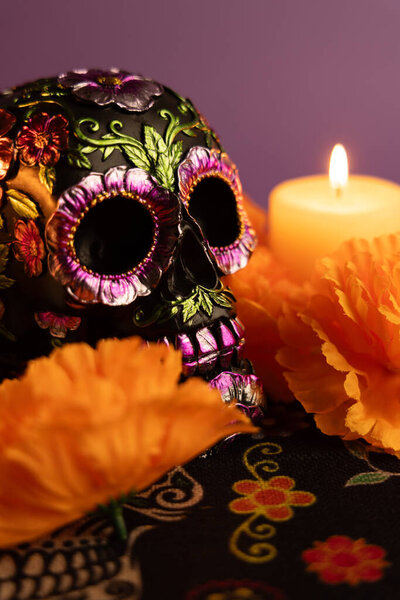 Красиво украшенный череп с металлическими цветами расположен в центре вертикального кадра. Одна зажженная свеча и окружающие оранжевые цветы напоминают праздник Диа-де-лос-Муэртос, когда устраиваются новые свечи..