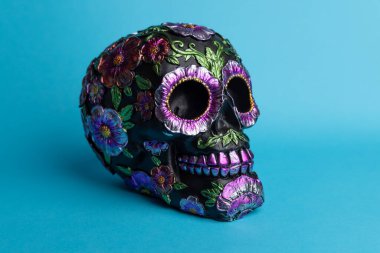 Metalik çiçeklerle bezenmiş siyah bir kafatasının çarpıcı bir görüntüsü, canlı mavi bir arka plan üzerine kurulmuş. Tasarım Meksika Ölüler Günü 'nü kutluyor. Karmaşık detaylar ve şenlikli renkler vurgulanıyor..