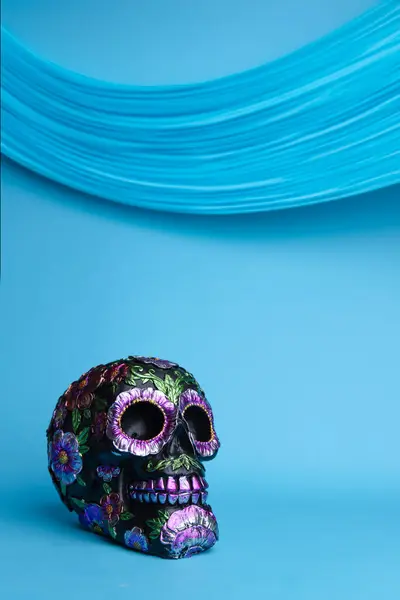 stock image A vertical shot of a black skull adorned with metallic flowers, set against a vibrant blue background. Perfectly captures the essence of Dia de los Muertos with a modern twist.