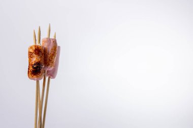 Sol tarafında kızarmış marşmelov olan şiş şişlerin yakın plan beyaz bir arka planı var. Fotoğraf, şekerlemelerin yatay bir düzende altın-kahverengi dokusunu vurguluyor.