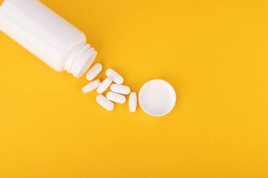 Beyaz bir ilaç şişesi beyaz hapları sarı zemine döküyor. Görüntü yukarıdan alındı, sağlık ve ilaç görselleri için uygun.