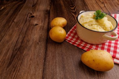 Bir kase kremalı patates püresi kahverengi ahşap bir masada oturur, yanında da tam patates ve kırmızı bir peçete. Rahatlatıcı yemek.