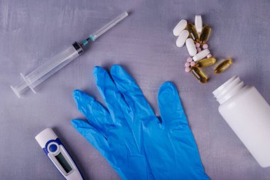 Üstten bakıldığında mavi bir nitril eldiven, termometre, şırınga, ilaç şişeleri ve bir defter görülüyor. Hepsi de beton bir yüzeye düzgünce yerleştirilmiş..