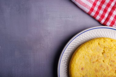 Lezzetli bir İspanyol patates omletinin mavi beton bir masada kırmızı kareli bir peçetenin yanında dinlenmesinin kısmi görüntüsü. Sıcak bir yemek sahnesi.