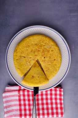 Lezzetli bir İspanyol patates omleti, bıçakla kesilmiş ve kırmızı kareli peçeteyle servis edilmiş, mavi beton bir masada.