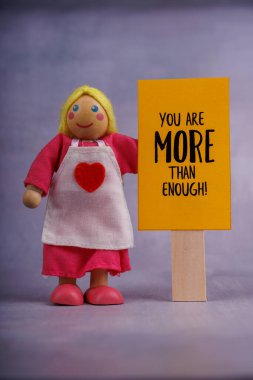 Tahta bir bebek, minimalist bir ortamda pozitiflik ve ilham mesajı veren turuncu bir motivasyon işareti taşır.