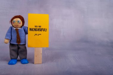 Tahta oyuncak figürü, basit ve oyuncu tasarımı sayesinde iyimserlik ve motivasyon saçan olumlu bir mesajla sarı bir işaret tutar.