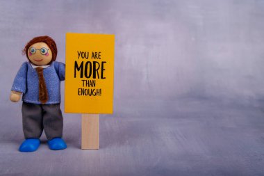 Tahta bir oyuncak figürü motive edici bir ifadeyle sarı bir işaret tutar. Sahne, neşeli ve minimalist bir şekilde pozitif enerji ve cesaret veriyor.