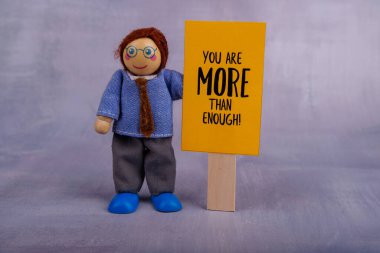 Tahta oyuncak figürünün elinde motivasyon mesajı taşıyan parlak sarı bir pankart var. Oyuncak bir sahnede pozitifliği ve teşviği simgeliyor.