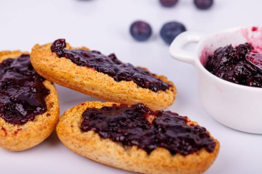 Böğürtlen reçelli bir dilim tost beyaz arka planda reçel kavanozunun yanında oturur. Taze ve canlı, bu kahvaltı sahnesi lezzet dolu.