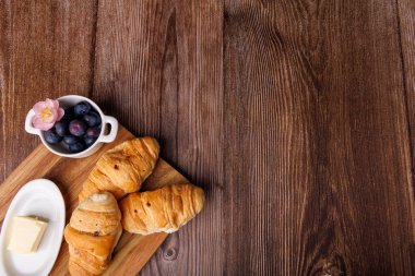 Ahşap bir kesim tahtasının yukarıdan görünüşü. Üzerinde yaban mersini reçeli ve koyu ahşap bir masada tereyağı kabı olan kruvasanlar var.