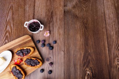 Yaban mersini reçelli tost ve ahşap bir tabakta bir tereyağı tabağı, koyu ahşap bir masaya kurulmuş, kahvaltı dokularını renklendiriyor.