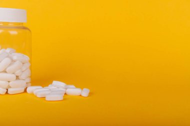 Sarı arka planda şeffaf haplar ve beyaz tabletlerle dolu açık bir şişe. Sağlık ve eczane malzemeleri sergileniyor.