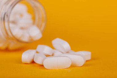 Sarı üzerine saçılmış tabletlerle birlikte açık ilaç şişesi, minimalist bir düzende sağlık ve tıbbı vurguluyor.