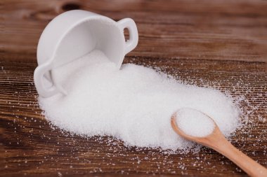 Bir seramik şeker kabı devrildi, tahtadan bir masanın üzerine şeker döküldü. Şeker dolu ahşap bir kaşık yakınlarda duruyor, kırsal ve doğal bir manzara yaratıyor.