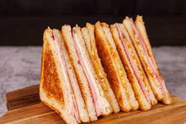 Jambonlu ve peynirli sandviçlerden oluşan büyüleyici bir düzenleme, ortadan ikiye bölünmüş ve siyah arkaplanlı koyu beton bir masada zarif bir şekilde sergileniyor.