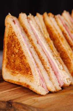 Tatmin edici bir jambonlu ve peynirli sandviç gösterisi. İkiye bölünmüş ve koyu beton bir masada siyah bir arka planda güzelce sunuluyor.