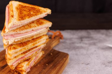 İkiye bölünmüş jambonlu ve peynirli sandviçlerden oluşan tatmin edici bir düzenleme. Gri beton bir masada, koyu siyah bir zemine karşı dikey olarak dizilmiş.