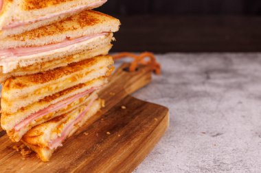 Dikey bir kule oluşturan canlı bir jambonlu ve peynirli sandviç gösterisi, grevdeki siyah arkaplanı olan gri beton bir masada ustalıkla düzenlenmiştir.