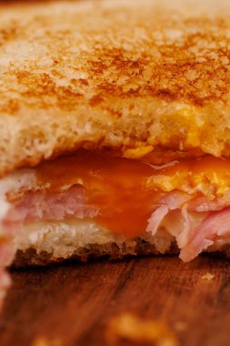 Canlı, yarısı yenmiş bir yumurta, jambon ve peynirli sandviç dikey bir kulenin tepesinde, koyu renk bir tahta üzerinde kırmızı vişneli domateslerle güzelce yerleştirilmiş.