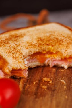 Dikey bir kulede yarısı yenmiş yumurta, jambon ve peynirli bir sandviç sergilenir ve yanında gri bir masada taze vişneli domatesler bulunur.