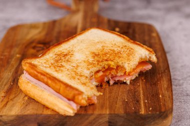 Tatmin edici yarısı yenmiş yumurta, jambon ve peynir sandviçi dikey bir kulede sergilenir, yanında gri bir masada taze vişneli domatesler.