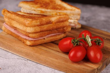 İkiye bölünmüş nefis bir jambonlu ve peynirli sandviç, koyu ahşap bir tahtanın üzerinde bir yığın taze vişneli domatesin yanında sergileniyor.