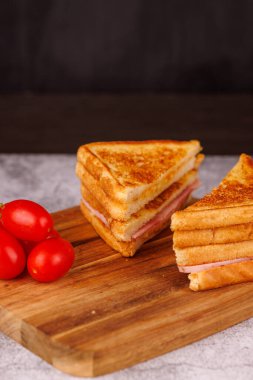 İkiye bölünmüş lezzetli bir jambonlu ve peynirli sandviç güzel bir şekilde koyu renk bir tahtada taze vişneli domatesle sunuluyor.