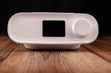 Görüntü, uyku apnesi tedavisinde kullanıldığını vurgulayan, koyu renk ahşap bir masaya yerleştirilmiş beyaz bir CPAP makinesini içeriyor.
