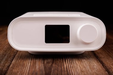 Ekran, hortum ve maskesiyle beyaz bir CPAP makinesi, koyu renk ahşap bir masada gösterilir. Uyku apnesi tedavisinde kullanıldığını vurgular.