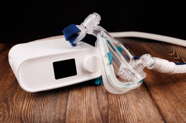 Görüntü, uyku apnesi tedavisinde kullanıldığını vurgulayan, koyu renk ahşap bir masaya yerleştirilmiş beyaz bir CPAP makinesini içeriyor.