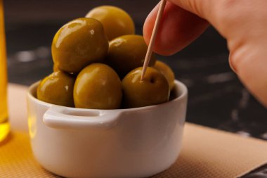 Bir el, kürdanlı bir zeytini bir şişe yağın ve siyah mermer masada bir tabak zeytinin yanına koyar.
